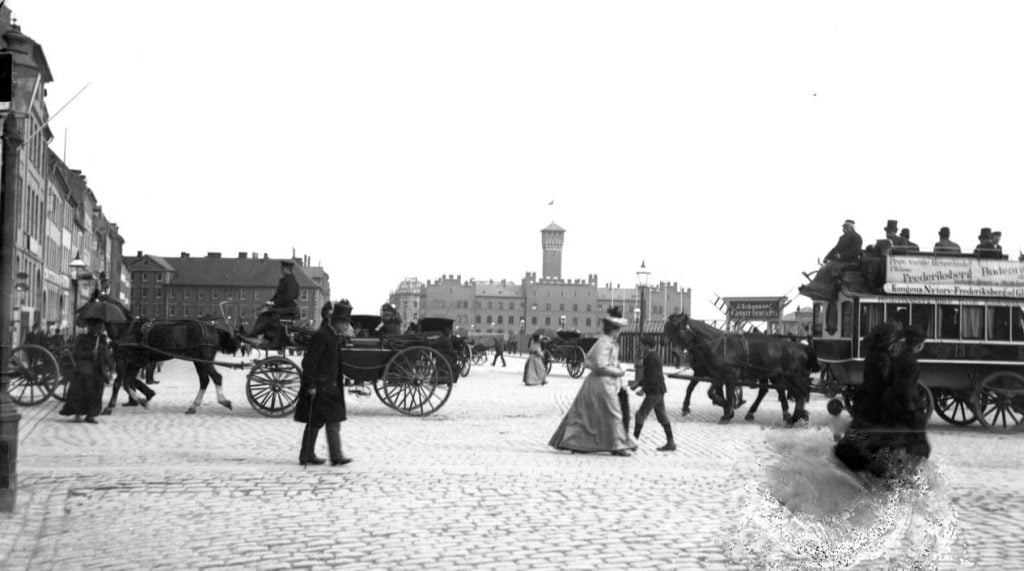 22.25 Rådhuspladsen (før 1893 Halmtorvet) set fra hjørnet af Vestergade mod Vartov. Brolagt plads med gående og kørende trafik, tv. en karet, th. en hestesporvogn (