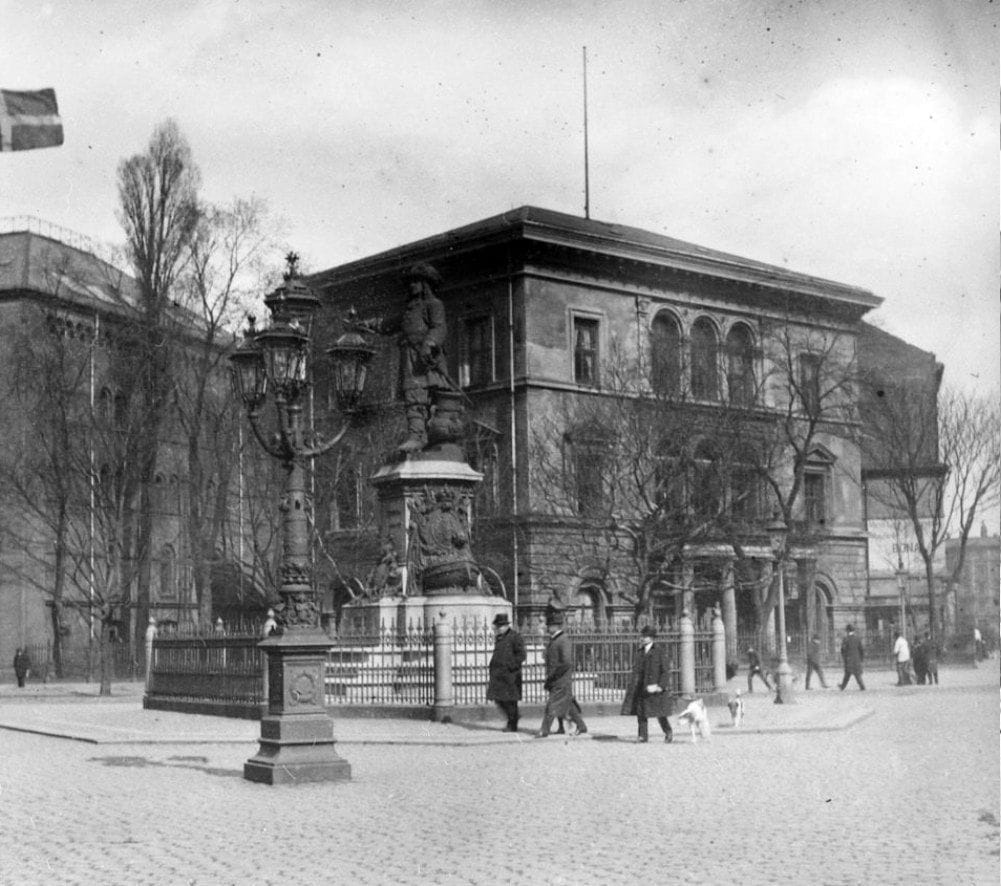 90.384 Holmens Kanal. Til højre i billedet ser vi ned ad den forholdsvis nyanlagte Holbergsgade. Den karakteristiske bygning på fotoet er den gamle Studenterforening, tegnet af J.D. Herholdt og opført i 1861-63 på resterne af Gammelholms gamle centrallagerpavillon. Foran bygningen står den store statue af Niels Juel.